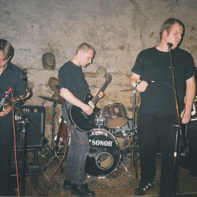 Vier Musiker vor einer Bruchsteinwand: Saxophon, Gitarre, Sänger am Mikrofon, zweite Gitarre